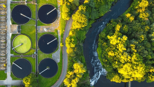 Aerial view of a wastewater treatment plant with circular sedimentation tanks next to a river and green forest