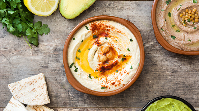 A bowl of creamy hummus topped with chickpeas, olive oil, paprika, and parsley, surrounded by pita bread, avocado, lemon, cilantro, and avocado dip on a rustic table
